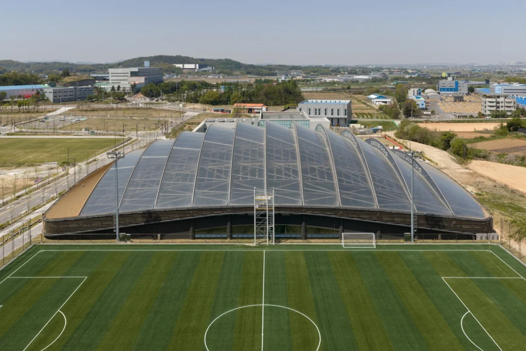 An elevated exterior shot of a large arched translucent roof structure next to a football pitch.