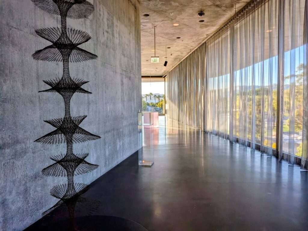 An interior gallery corridor with a textured concrete accent wall on the left and a continuous floor-to-ceiling glass wall with sheer curtains on the right.