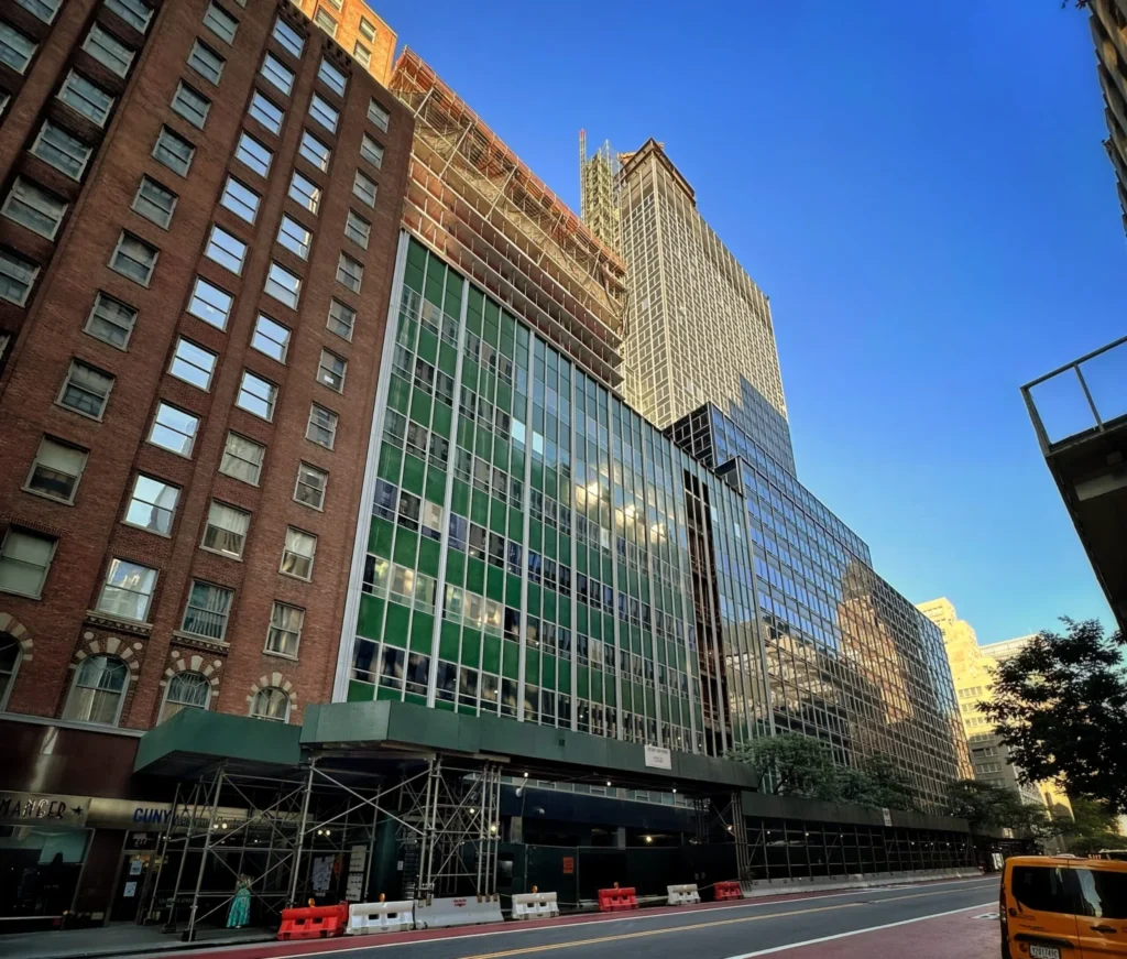 Street-level view of a building facade with green panel cladding and construction scaffolding along the sidewalk.