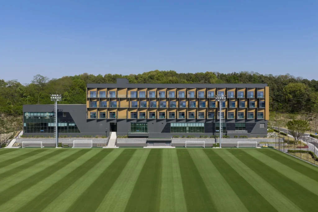 A direct facade view of a modern multi-story residential building featuring gold modules next to a green pitch.