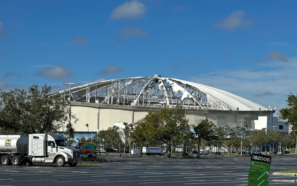 Exterior view of a domed stadium showing severe storm damage to its fabric roof paneling.