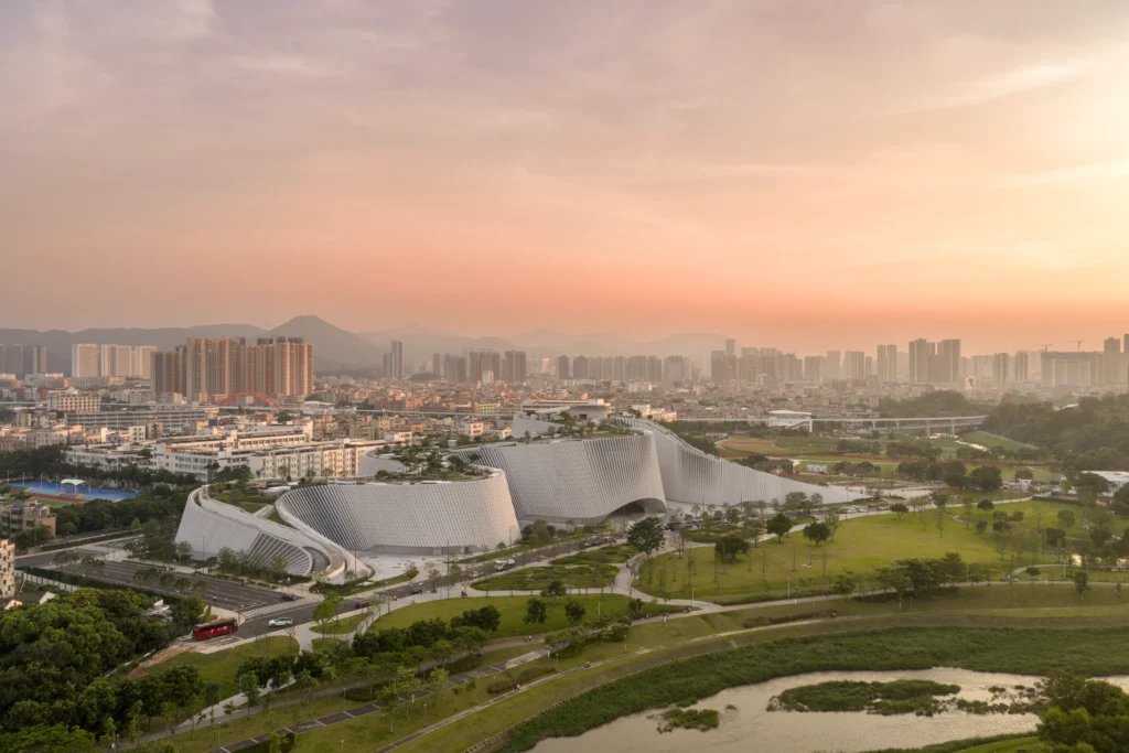 Aerial view of a long curving landform building with rooftop gardens adjacent to a river and dense city density.