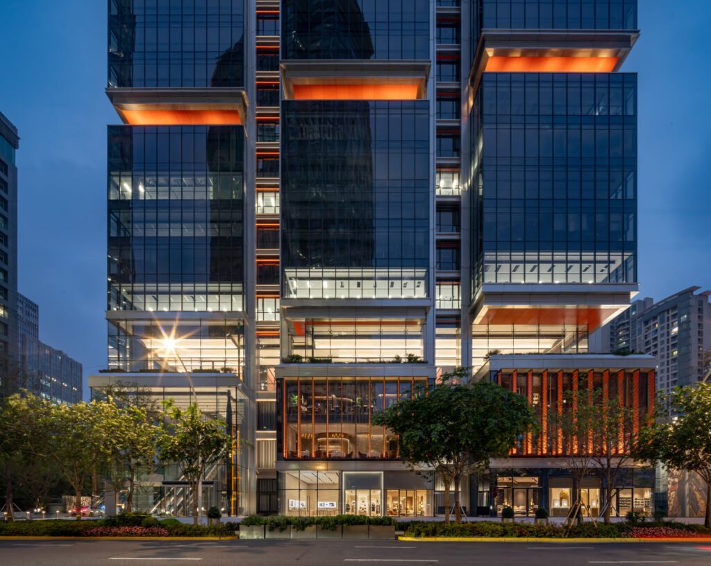 Night elevation view of illuminated base levels, orange louvered panels, and upper office modules.
