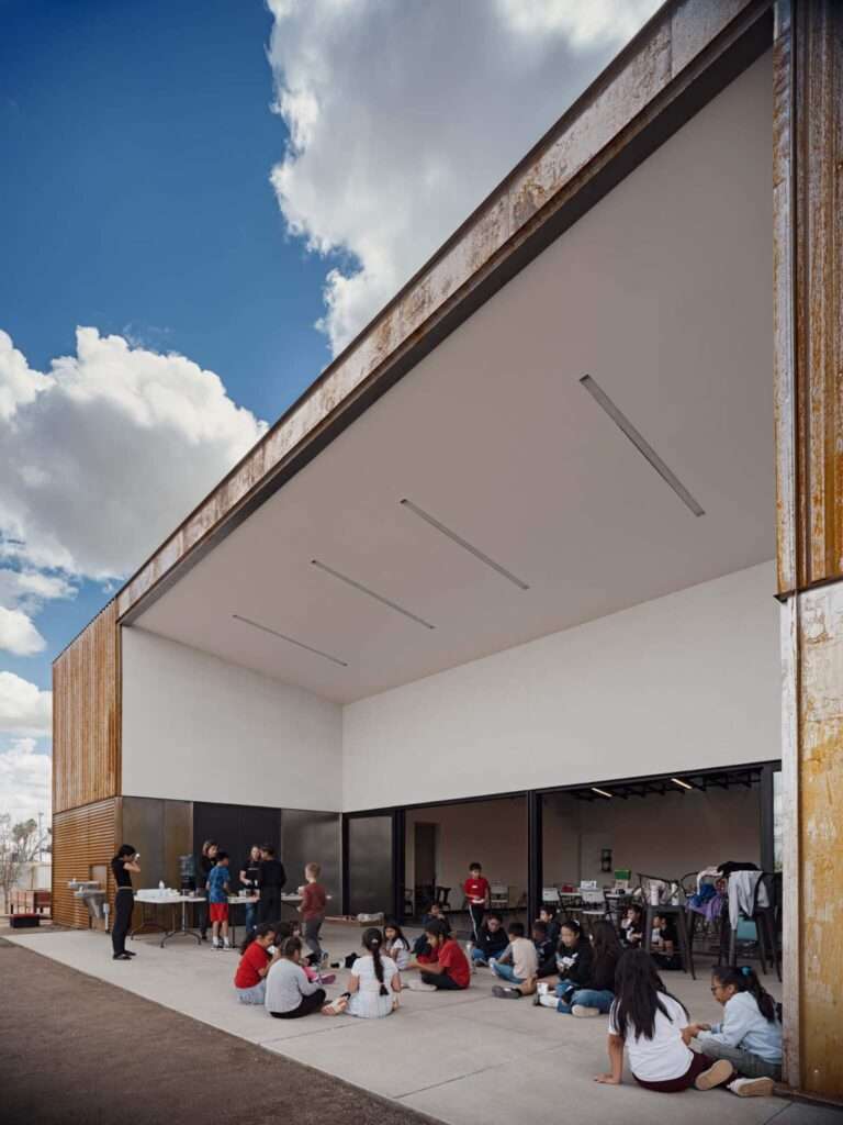 A building facade featuring extensive glass fenestration under a dark roof canopy, with a concrete walkway and masonry planters.