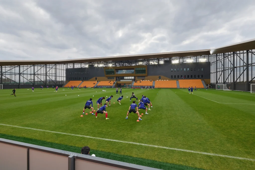 A group of football players in blue training kits stretching on a green field within the stadium structure.