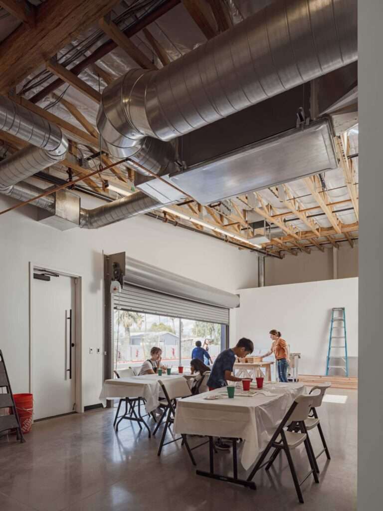 Children sitting on an outdoor concrete patio beneath a large cantilevered canopy clad in weathered metal.