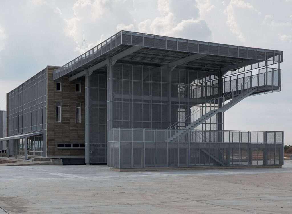 Exterior view of a multi-story industrial building featuring a textured concrete block core wrapped in a translucent steel mesh screen structure and an external staircase.