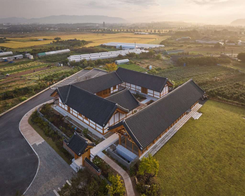 Aerial drone view of the JK-AR architectural project in Jeonju, showing three interconnected Hanok-inspired timber pavilions arranged around traditional Madang courtyards surrounded by agricultural fields at sunset.