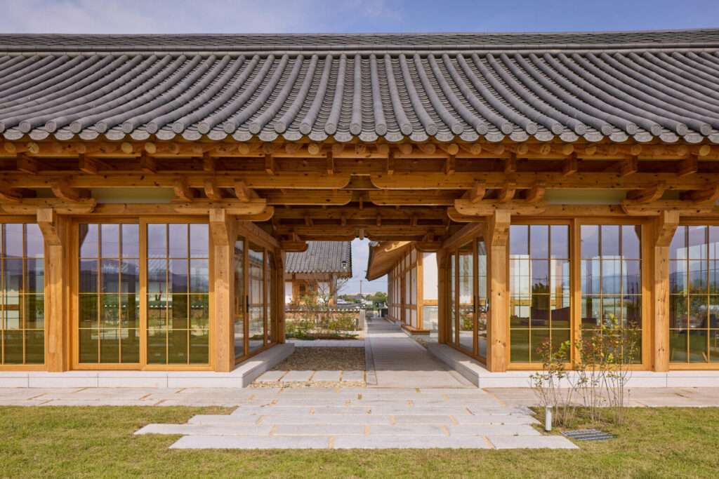 Ground-level view looking through an open-air timber breezeway under a dark tiled roof, showing the Toenmaru transition space and glazed wooden facades.