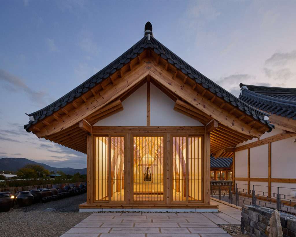 Exterior evening view of a gabled timber pavilion with full-height glass windows radiating warm interior lighting, with traditional ceramic pots lined on the left.