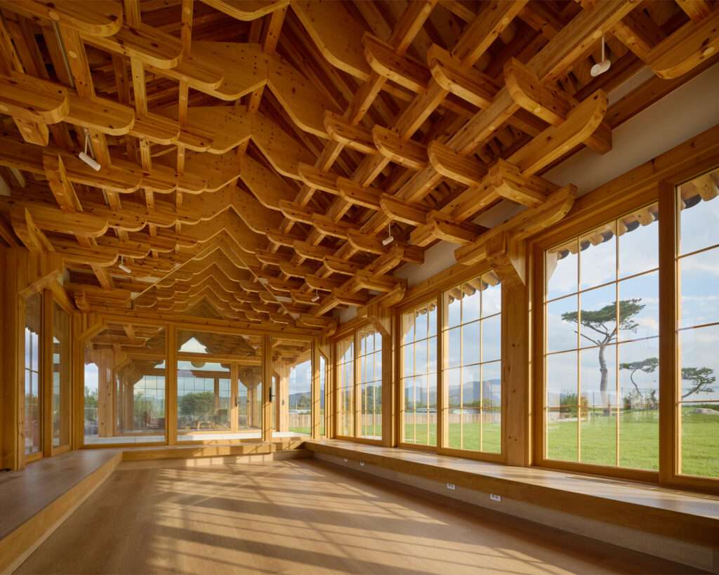 Interior view of the tea house pavilion featuring an intricate, dynamic wooden bracket ceiling system and large glass windows overlooking a green grass lawn.