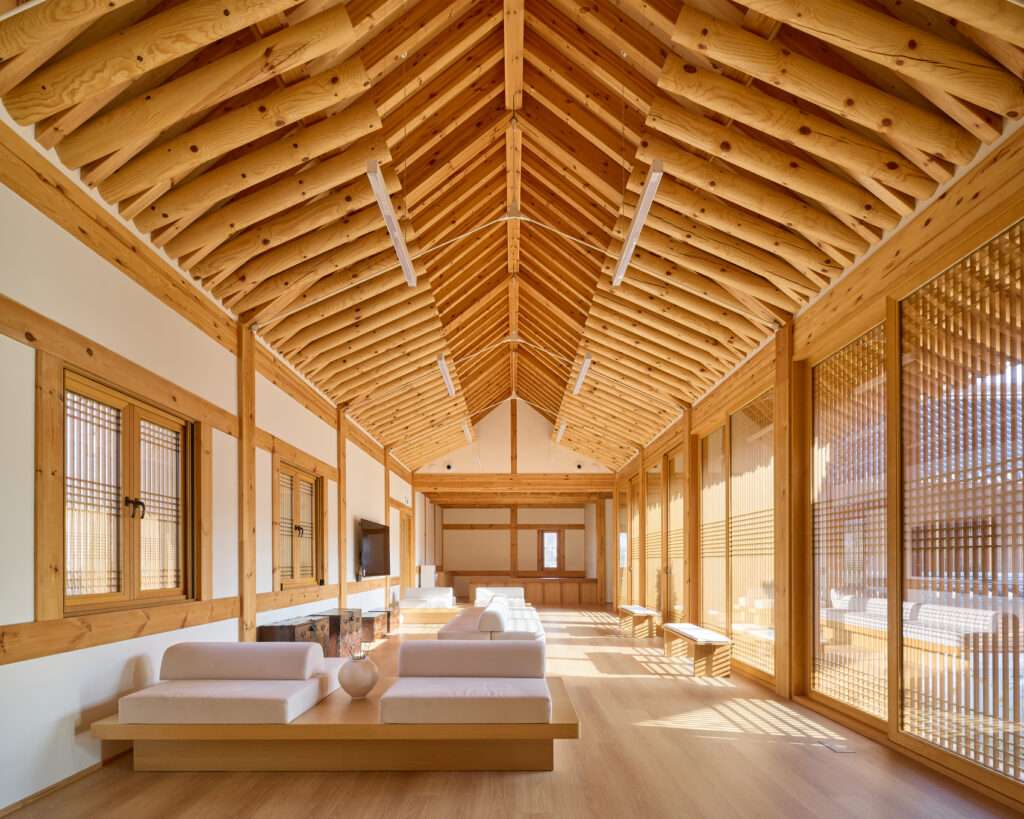 Interior view of a long, minimalist waiting lounge with light wood floors, built-in low sofas, and a exposed timber log rafter ceiling with vertical wooden screen panels.