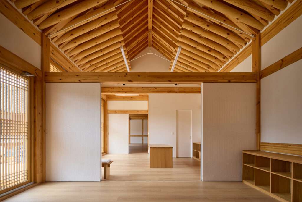 Minimalist interior space showing white sliding partition walls under an exposed heavy timber roof structure with built-in wooden shelving units.