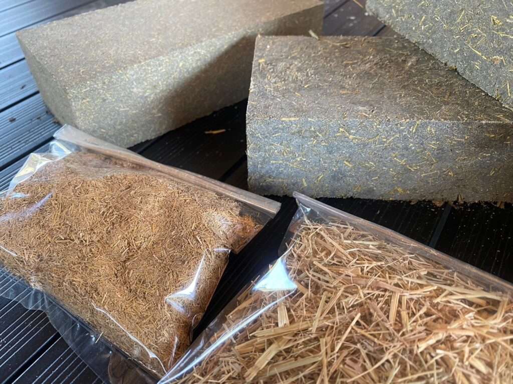 Raw agricultural waste samples including shredded rice straw and husks in plastic bags placed next to finished sustainable concrete masonry blocks.