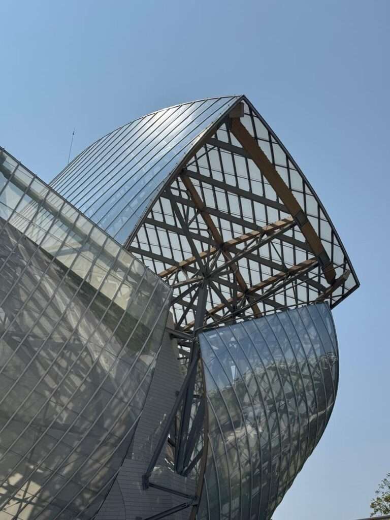 Looking up at the complex exterior architecture featuring sweeping, curved glass sails supported by a dense network of wooden beams and steel columns against a clear blue sky.