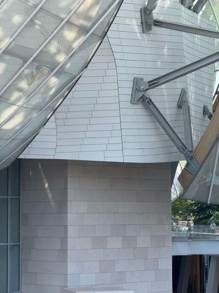Detailed view of the building's exterior showing a transition from a solid, curved white-paneled facade above to a sturdy, light-colored stone block base below, intersected by heavy steel support struts holding a glass canopy.