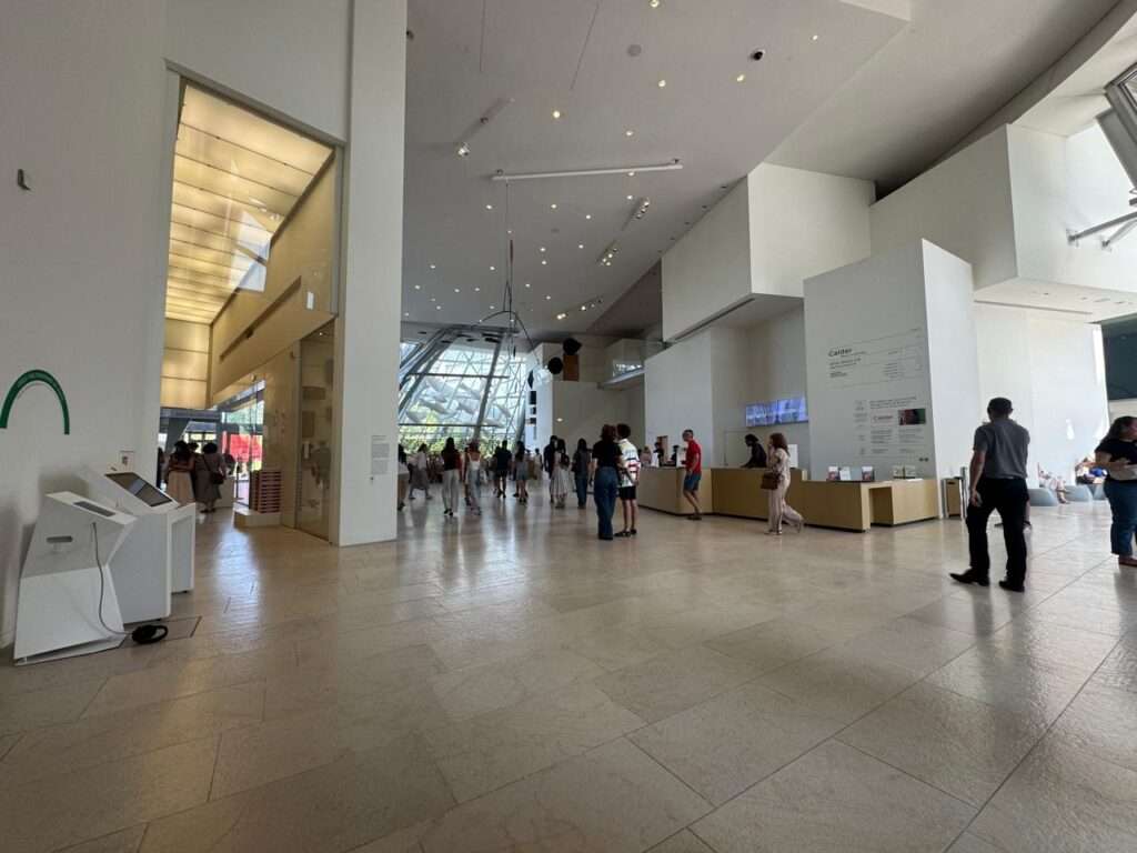 Wide interior view of the brightly lit main lobby, featuring white walls, a polished stone floor, visitor reception desks, and people walking, with a view of the glass architecture visible through a large window.