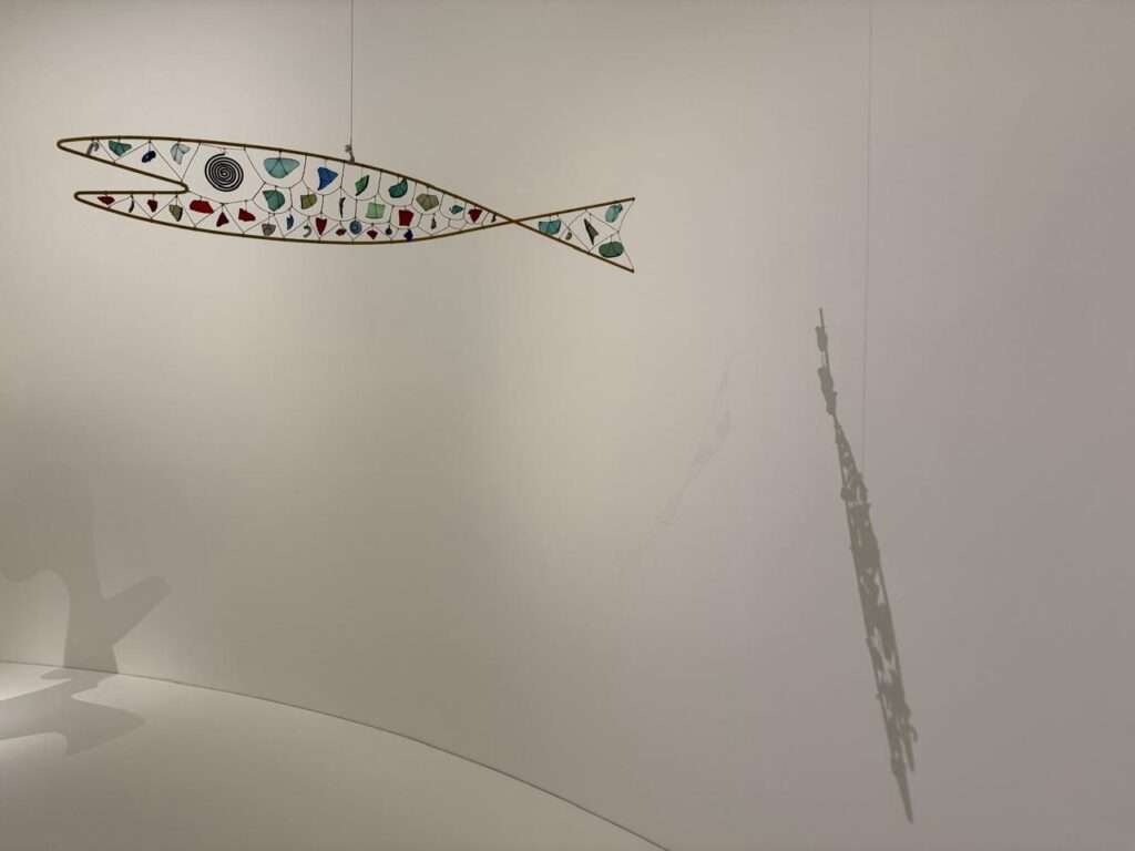 A delicate, fish-shaped kinetic mobile sculpture made of wire and colored glass pieces suspended against a curved white gallery wall, casting a distinct shadow to the right.