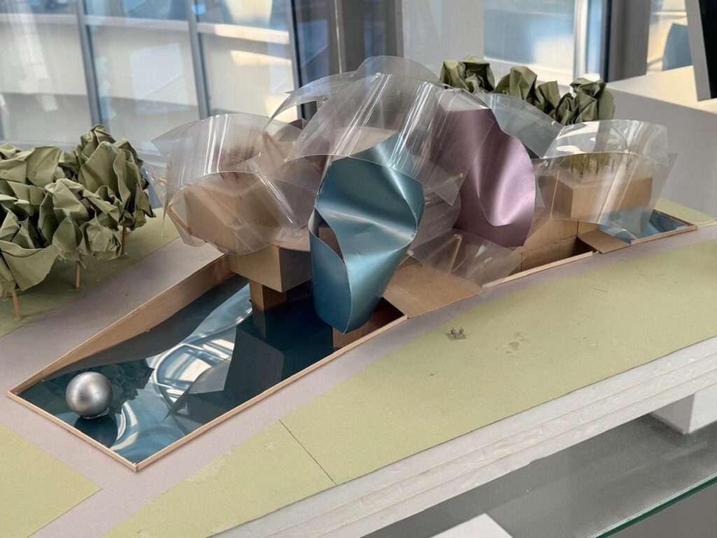 An architectural scale model of a deconstructivist building featuring dynamic, curved transparent and metallic-colored elements resembling sails, set next to a reflective pool and stylized paper trees.