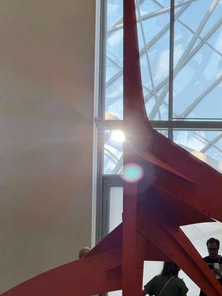 Close-up of a massive red steel structural support beam inside a modern museum, with bright sunlight creating a glare through a complex glass and steel roof structure in the background.