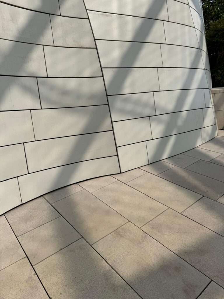 Close-up of a curved exterior wall clad in elongated white rectangular panels, highlighting a prominent vertical seam where two curving surfaces meet, casting sharp diagonal shadows.