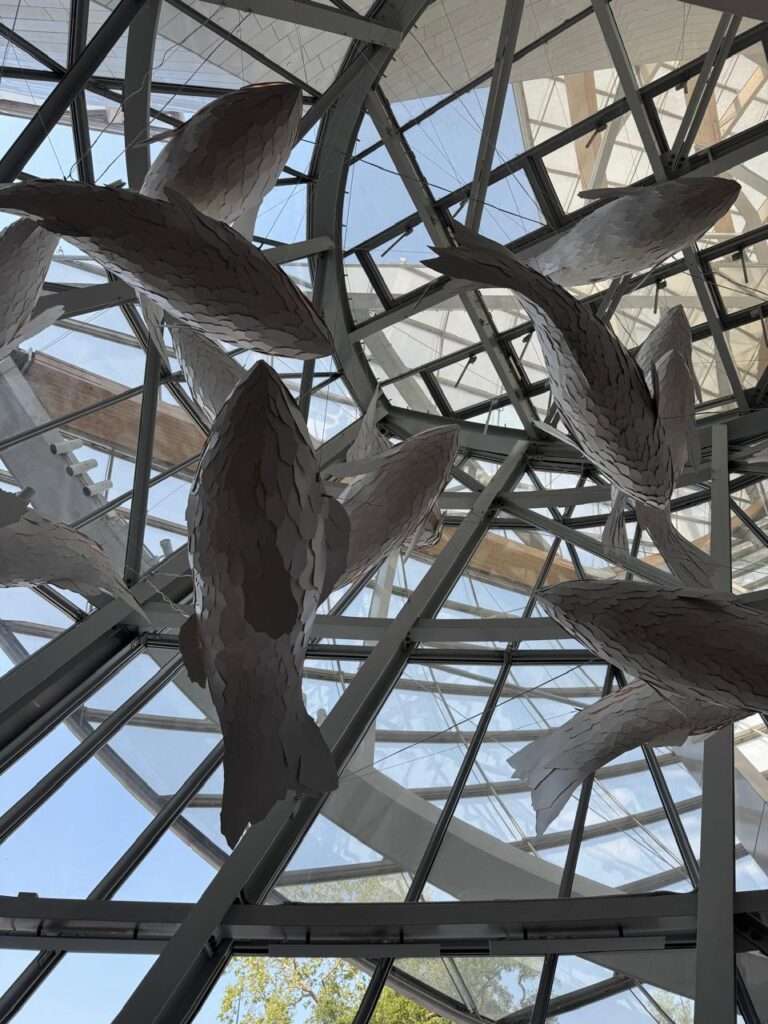 Low-angle view looking up at large, stylized fish sculptures suspended beneath an intricate ceiling made of intersecting steel beams and translucent glass panels.