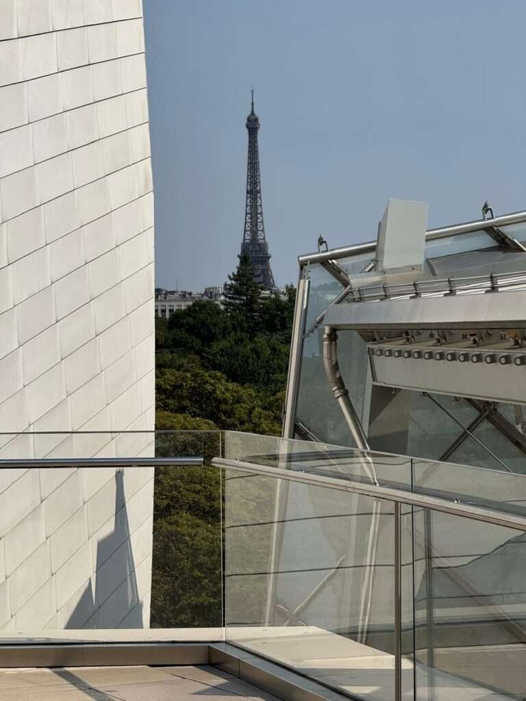 View of the Eiffel Tower rising above a dense green forest, framed by a smooth white paneled wall on the left and a complex glass and steel canopy structure on the right.