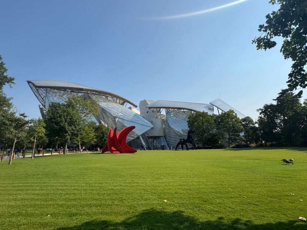 The Fondation Louis Vuitton building with its curved glass sails, seen from across a green lawn with a red sculpture
