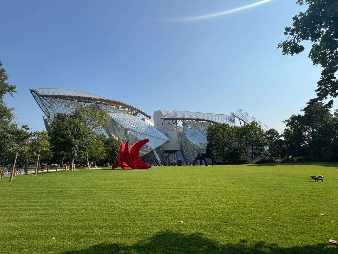 The Fondation Louis Vuitton building with its curved glass sails, seen from across a green lawn with a red sculpture