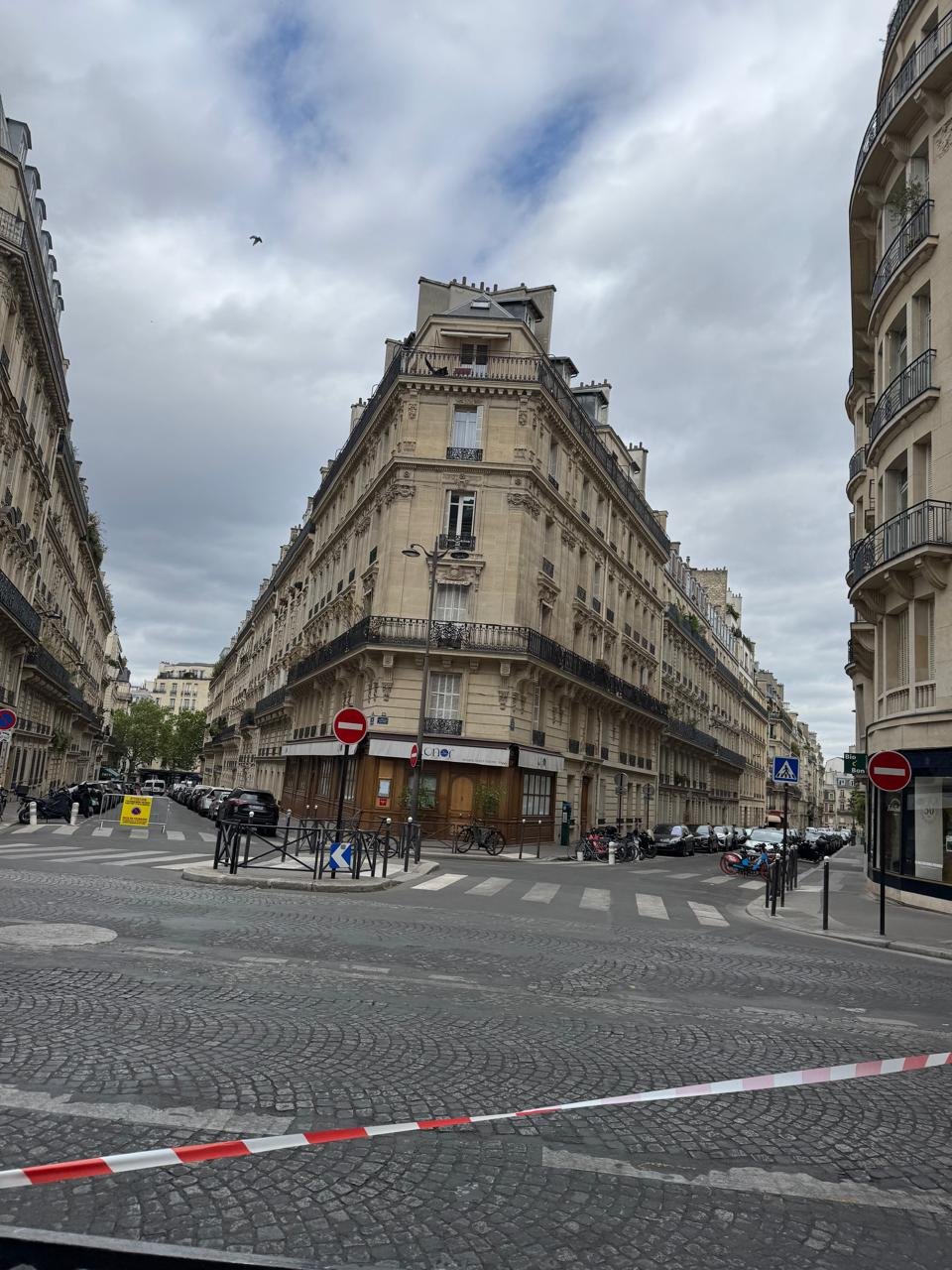 Red and white barrier tape blocking a typical Parisian intersection lined with traditional Haussmann residential buildings and ground-floor cafes under a cloudy sky.