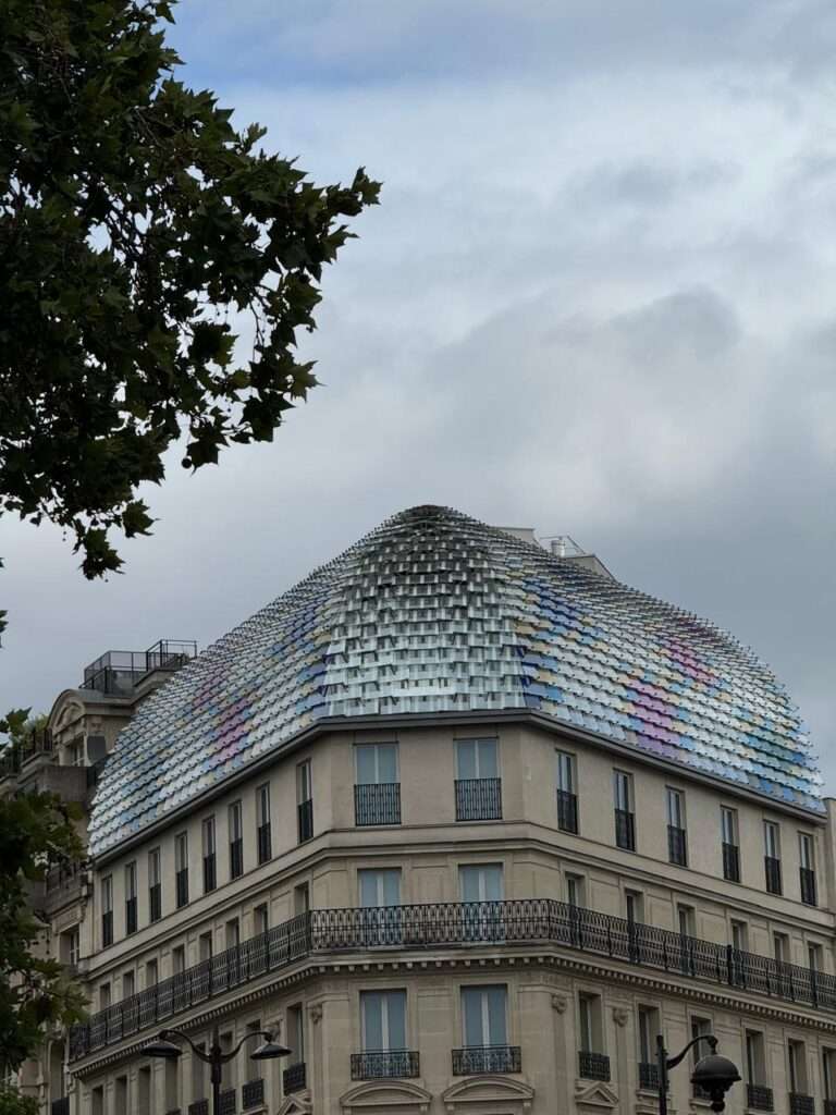 A contemporary, iridescent scaled glass dome sitting atop a traditional stone Parisian building against an overcast sky.