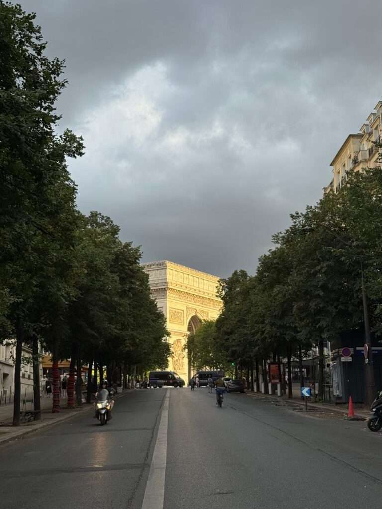 A view down a tree-lined Parisian avenue toward the sunlit Arc de Triomphe under a stormy sky, with official vehicles blocking routine vehicular traffic.