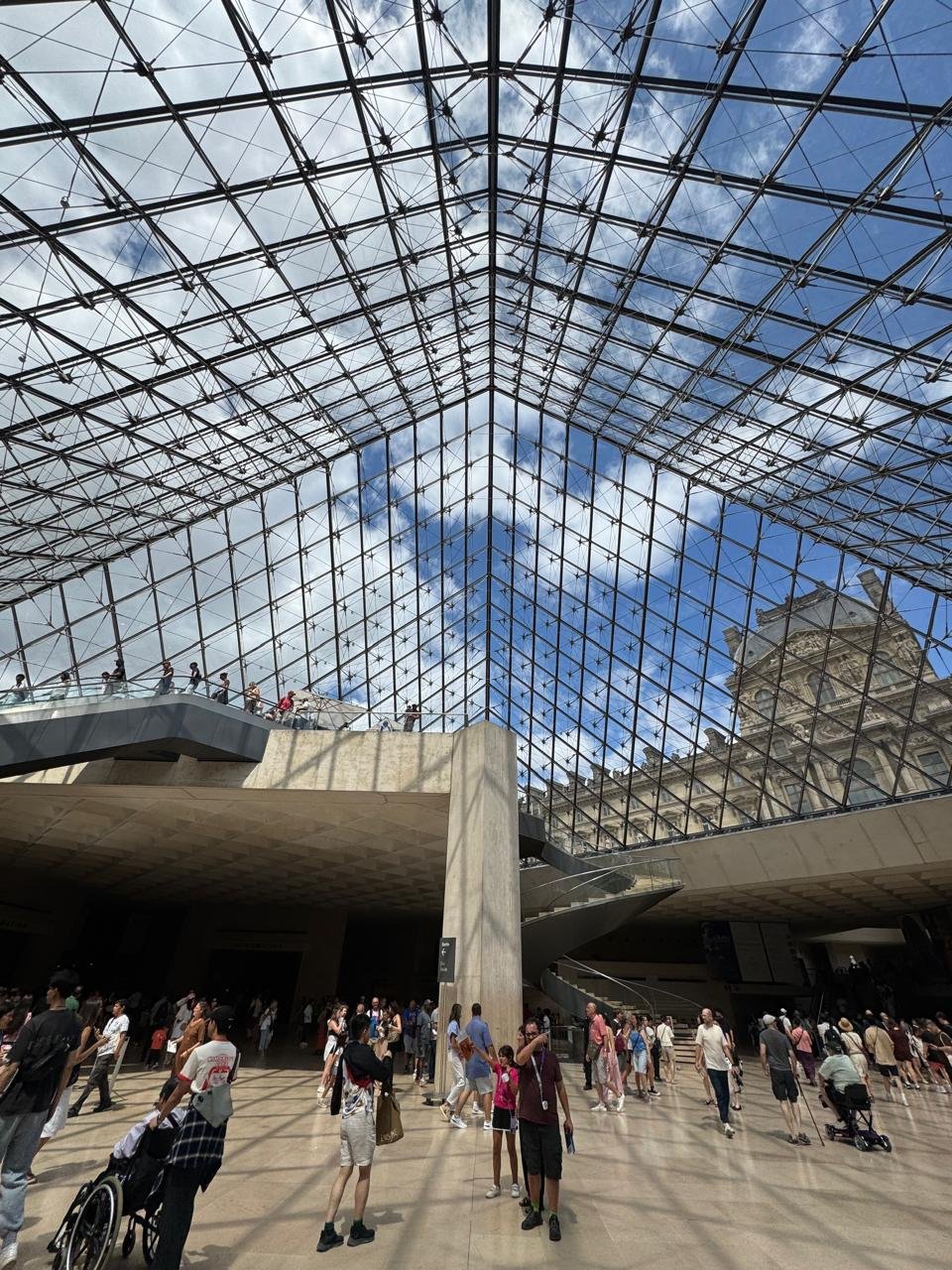 Interior view looking up through the geometric steel and glass structure of the Louvre Pyramid, with the historic museum facade visible in the background and visitors in the lobby below.