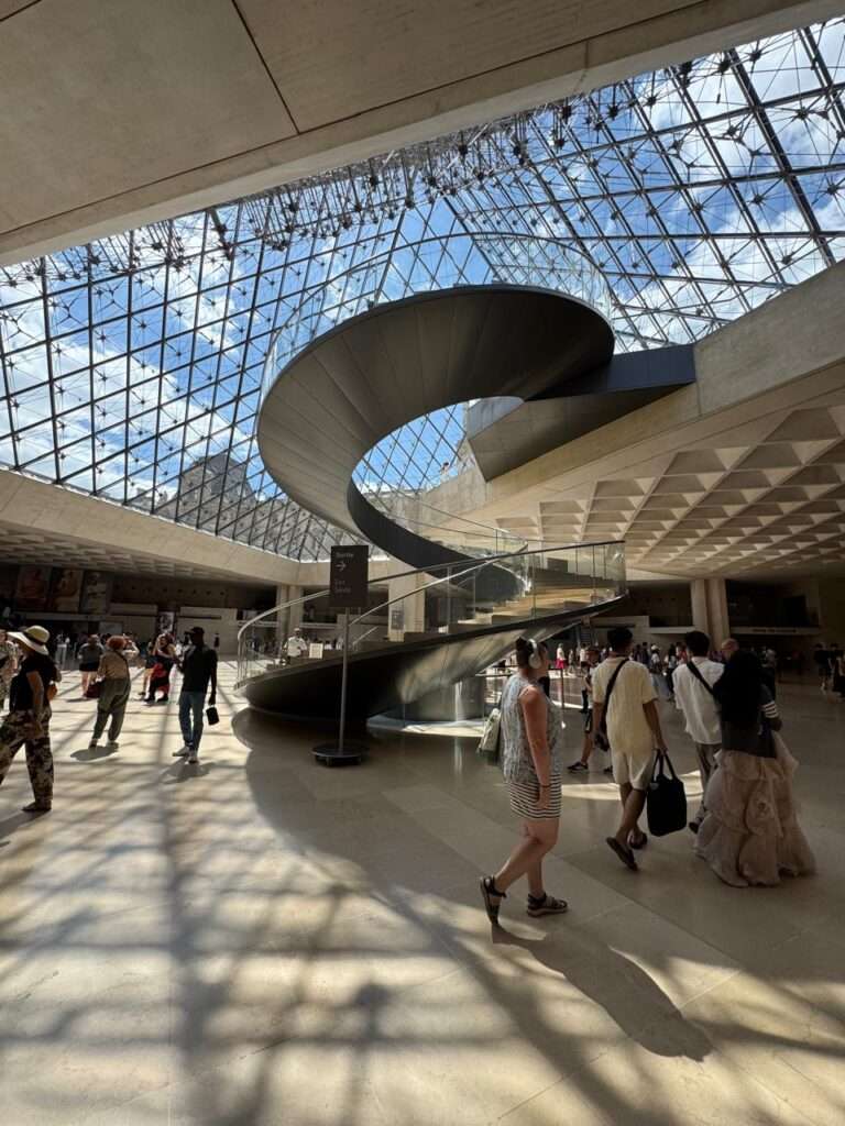 A sleek, modern helical staircase winding upward in the bright, underground lobby beneath the geometric glass roof of the Louvre Pyramid.