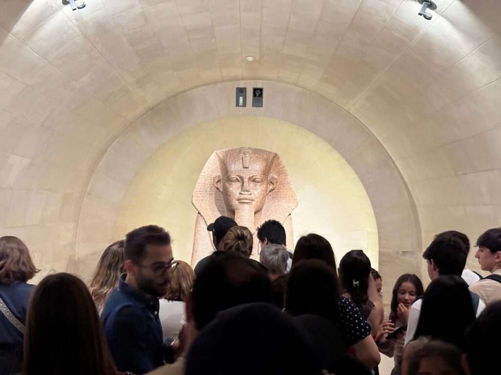 A massive Egyptian stone sphinx positioned at the end of a long, dimly lit barrel-vaulted stone corridor, surrounded by a crowd of onlookers.