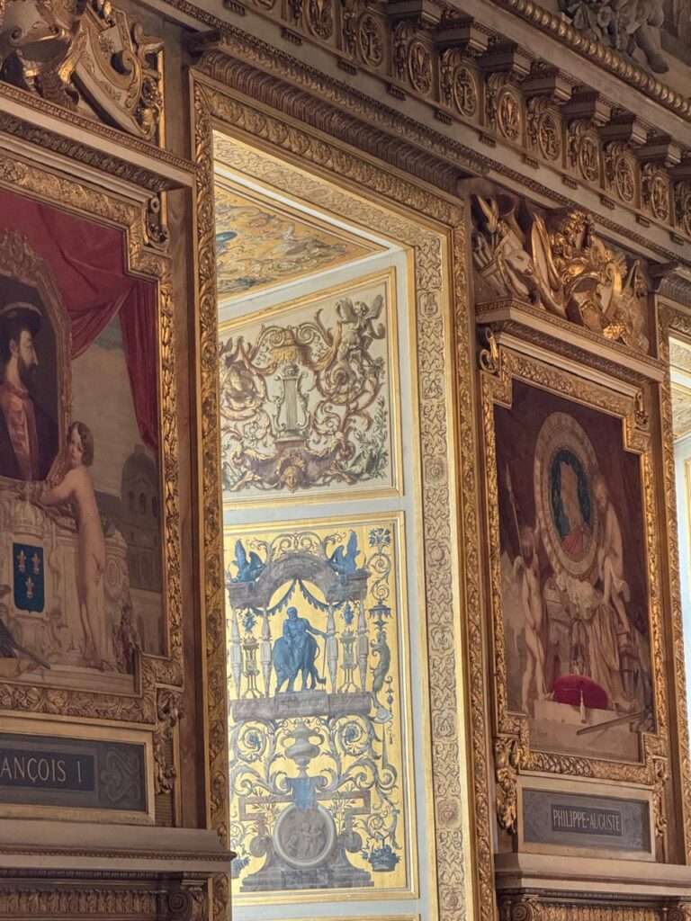 Close-up of ornate gold architectural frames enclosing classical portraits on a gallery wall, with a glimpse into a brightly lit adjacent room featuring blue and gold decorative motifs.