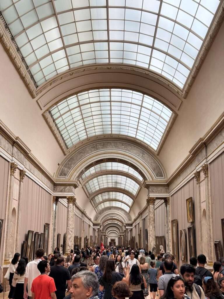 A remarkably long museum gallery with a massive glass skylight running along its barrel-vaulted ceiling, walls lined with paintings, and a dense crowd of visitors.