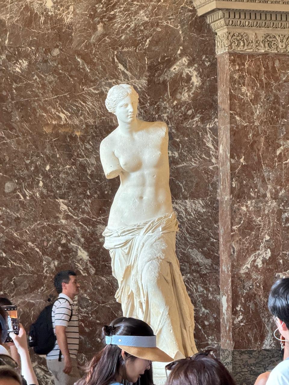 Venus de Milo displayed against veined red marble at the Louvre, with visitors visible around the sculpture.