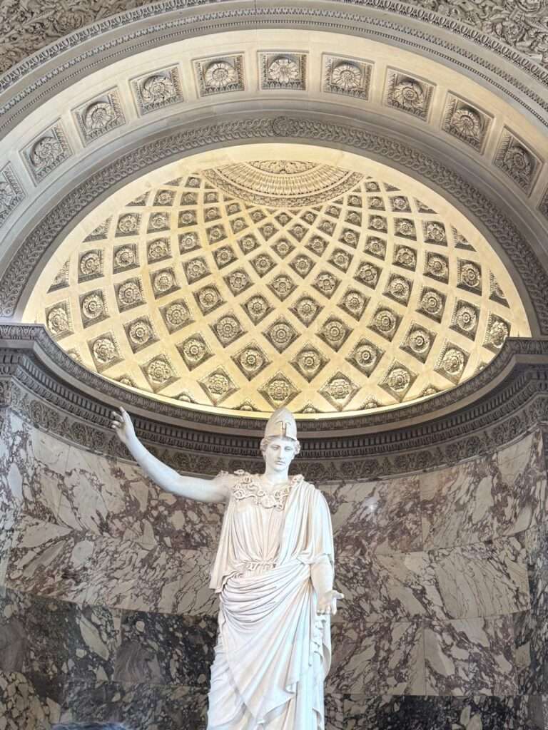 A towering classical marble statue of a helmeted figure standing in an architectural niche beneath a highly detailed coffered half-dome ceiling.