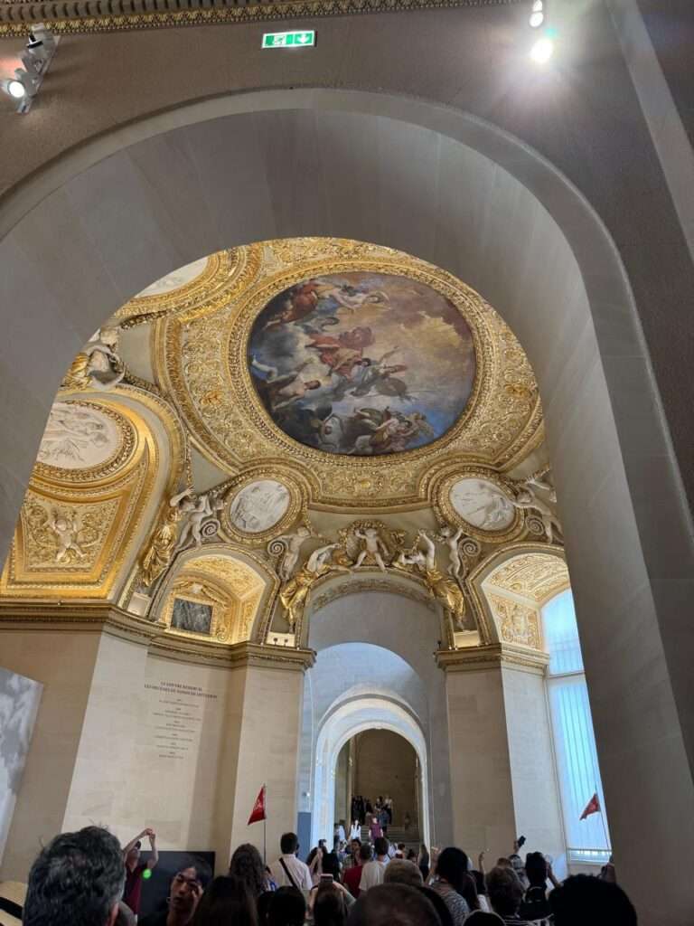 A highly decorated architectural transition space featuring a domed ceiling with a central fresco, surrounded by ornate gold relief sculptures and classical white medallions.