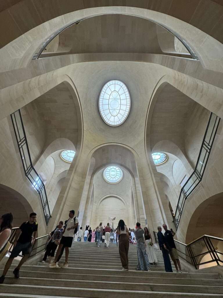 Looking up a monumental stone staircase beneath sweeping arches and domed skylights, with the Winged Victory of Samothrace visible at the top landing.