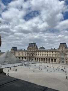 Elevated view of the Louvre Museum courtyard showing the main glass pyramid and smaller pyramids surrounded by classical stone facades under a cloudy sky.