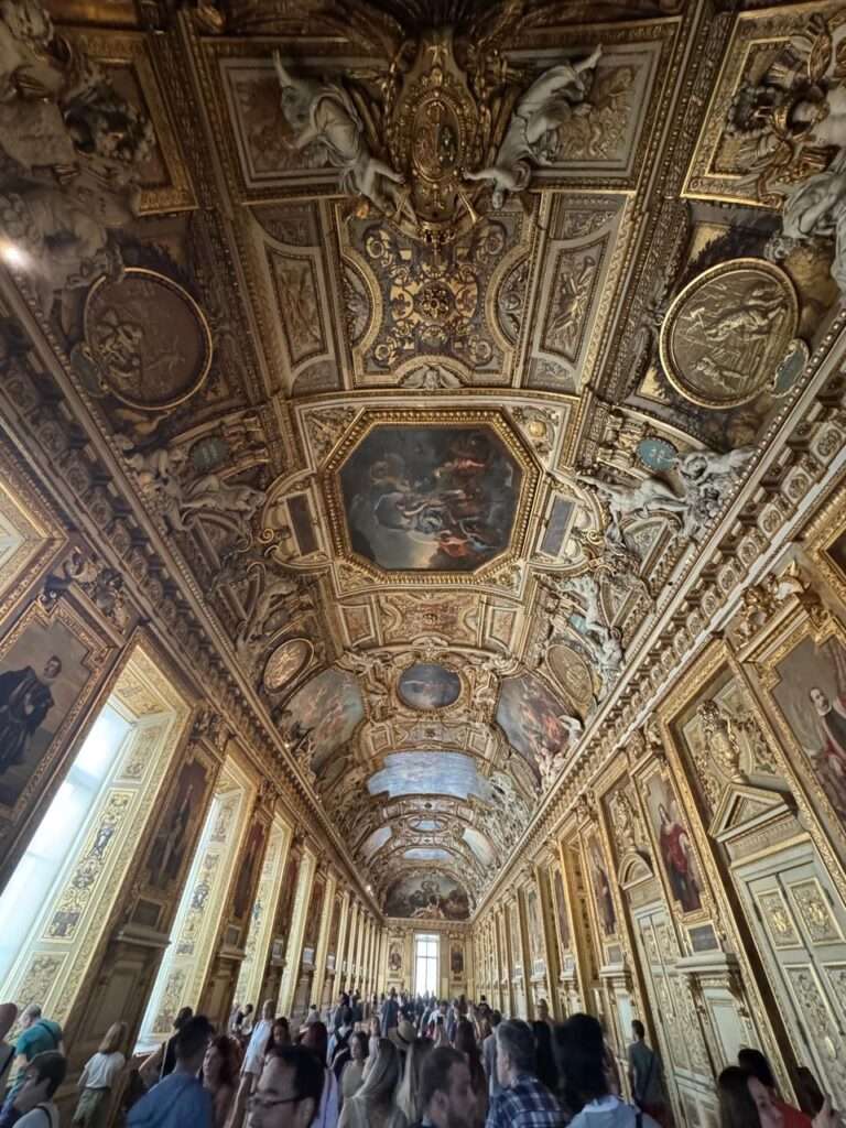 A long, palatial gallery with a magnificent vaulted ceiling covered in intricate gold stucco and inset paintings, filled with crowds of tourists.