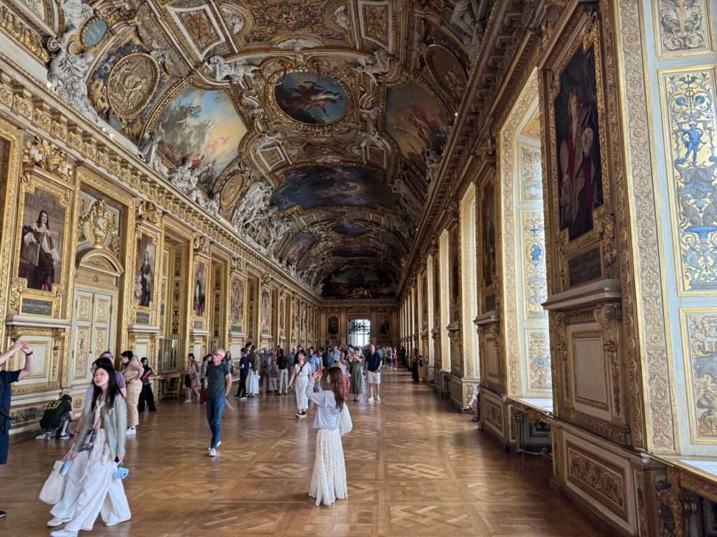 Wide view of a highly ornate historic gallery featuring a gilded vaulted ceiling, walls lined with framed portraits, tall windows, and intricate wooden parquet flooring.