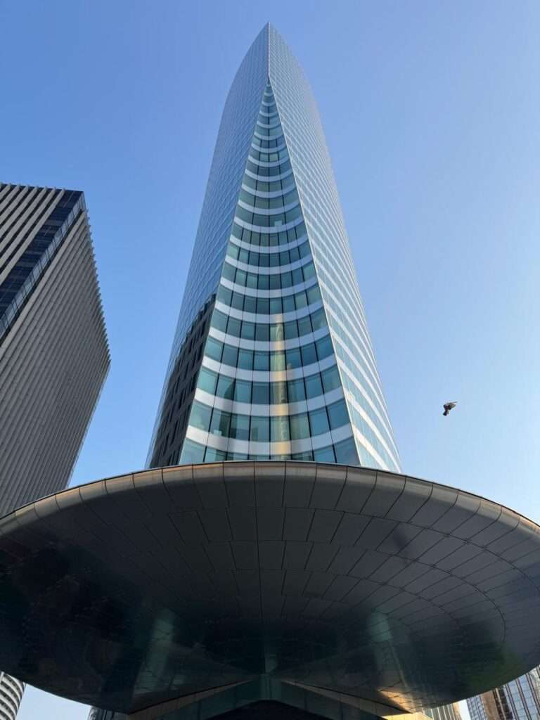 A dramatic low-angle view looking straight up the curved glass facade of a modern skyscraper, featuring a prominent, circular metal canopy at its base against a clear blue sky.