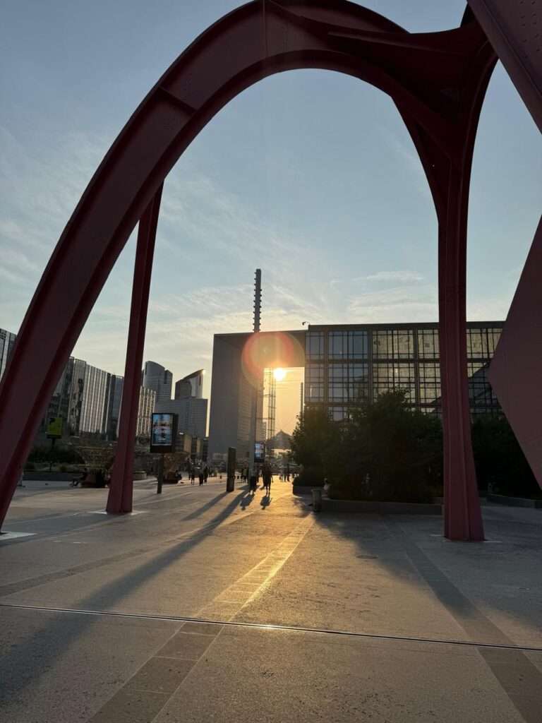 Sunset viewed through the massive legs of a monumental red steel sculpture, casting long, dramatic shadows across a textured concrete pedestrian plaza towards the Grande Arche de la Défense and surrounding glass skyscrapers.