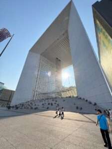 Low-angle view of the massive white Grande Arche de la Défense bathed in bright sunlight, with crowds of diverse people sitting on its wide monumental steps and walking across the textured concrete plaza.