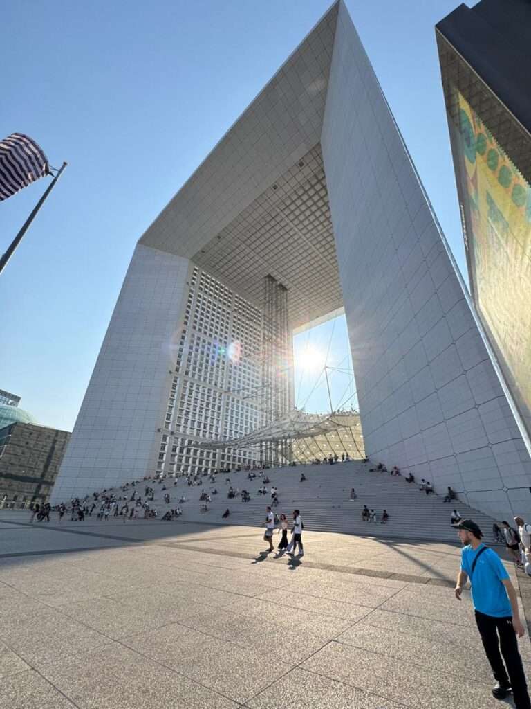 Low-angle view of the massive white Grande Arche de la Défense bathed in bright sunlight, with crowds of diverse people sitting on its wide monumental steps and walking across the textured concrete plaza.