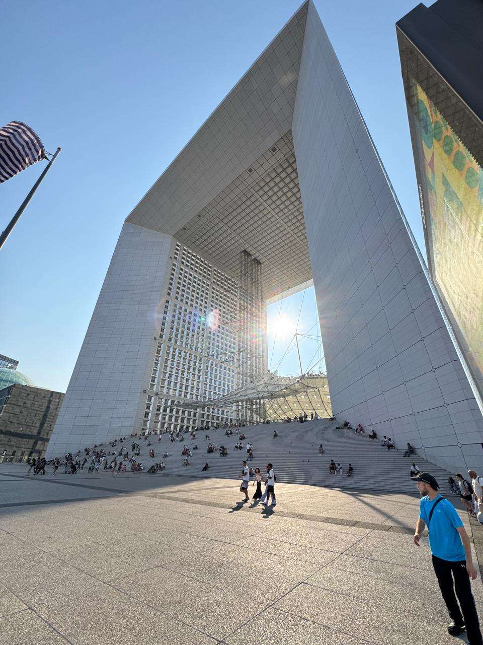Low-angle view of the massive white Grande Arche de la Défense bathed in bright sunlight, with crowds of diverse people sitting on its wide monumental steps and walking across the textured concrete plaza.
