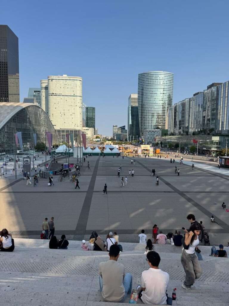 Elevated view from the concrete steps of the Grande Arche looking toward the La Défense esplanade. A highly diverse, casually dressed crowd lounges in the foreground, with an American-style branded beverage carton resting on the stairs. Temporary event tents and modern glass skyscrapers fill the background under a clear sky.